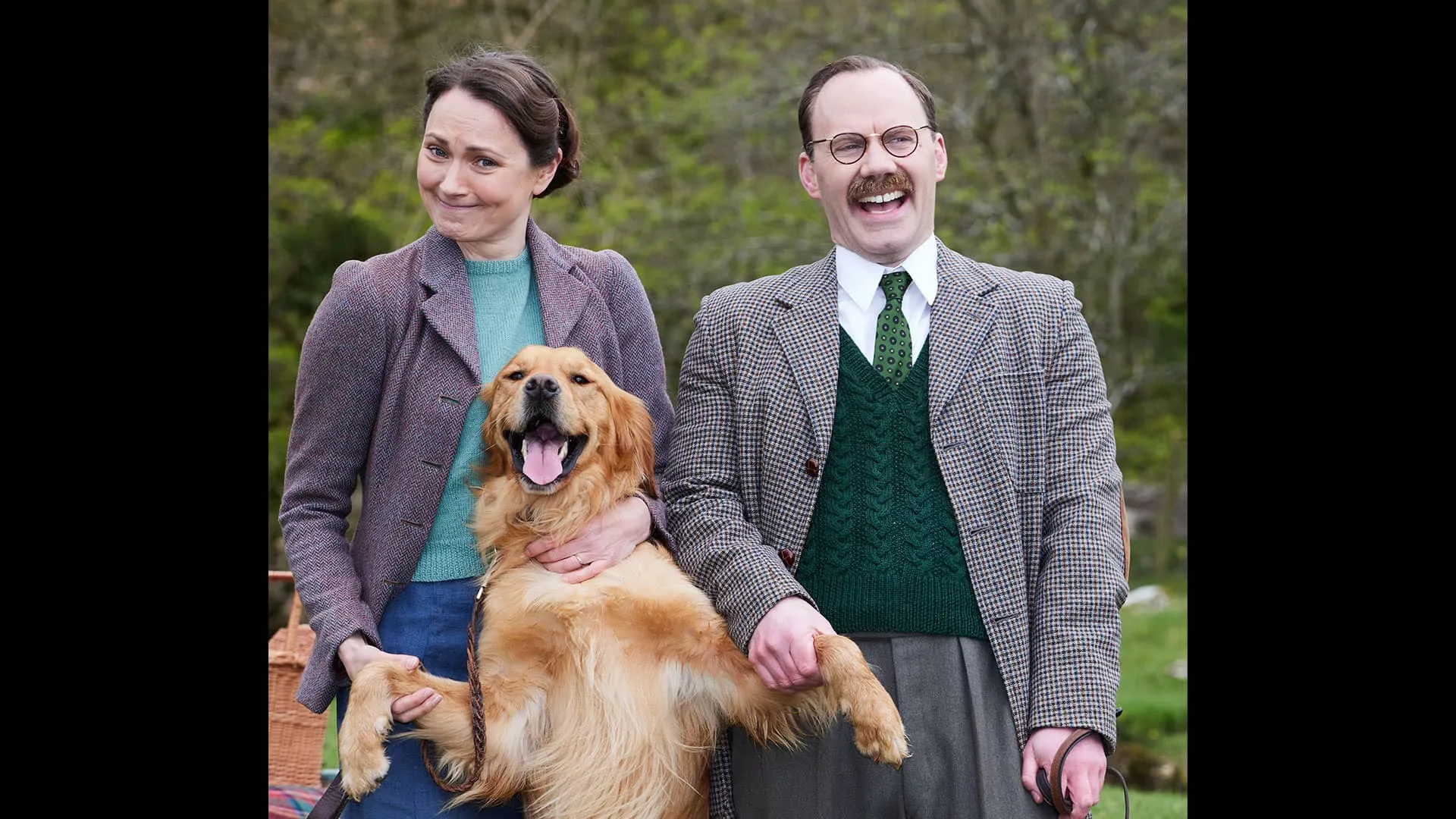 actress Anna Madeley (Mrs. Hall) and Will Thorpe (Gerald) tand outdoors with golden retriever Ernie (Jess) on his hind legs between them, looking happy with his tongue out. Will laughs and Anna smiles