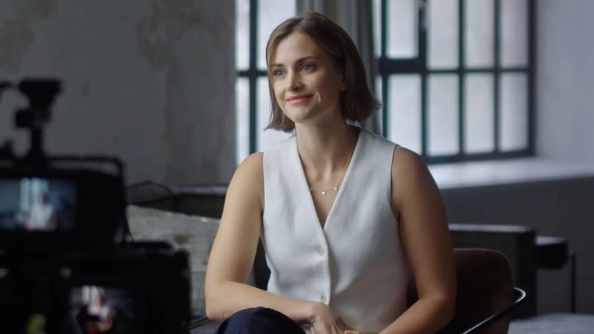 Actor Stefanie Martini, seated and wearing a white vest, smiles at an interviewer off camera.